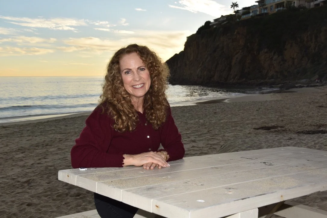 Parenting coach Jeanine Mouchawar sitting at a beachside table at sunset, leaning forward with a warm smile