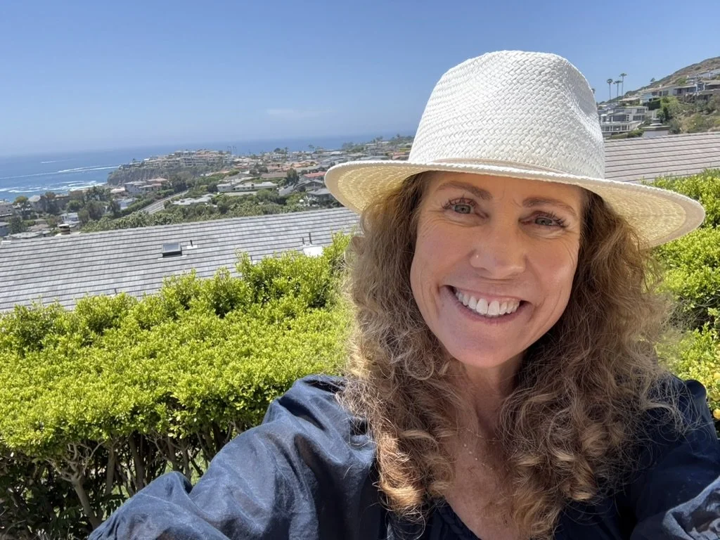 Jeanine Mouchawar smiling outdoors in a white sun hat overlooking the ocean, accompanying a blog about back-to-school parenting and knowing when to step in or step back.