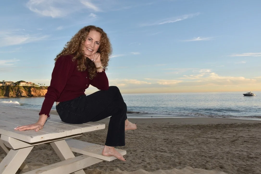 Parenting coach Jeanine Mouchawar sitting on a beach picnic table at sunset, chin resting on hand, looking directly at camera