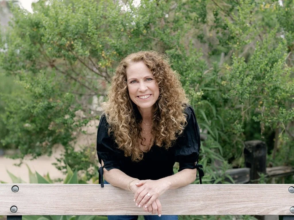 Jeanine Mouchawar, parenting coach, leaning forward on a wooden bench outdoors, smiling warmly in a calm natural setting.