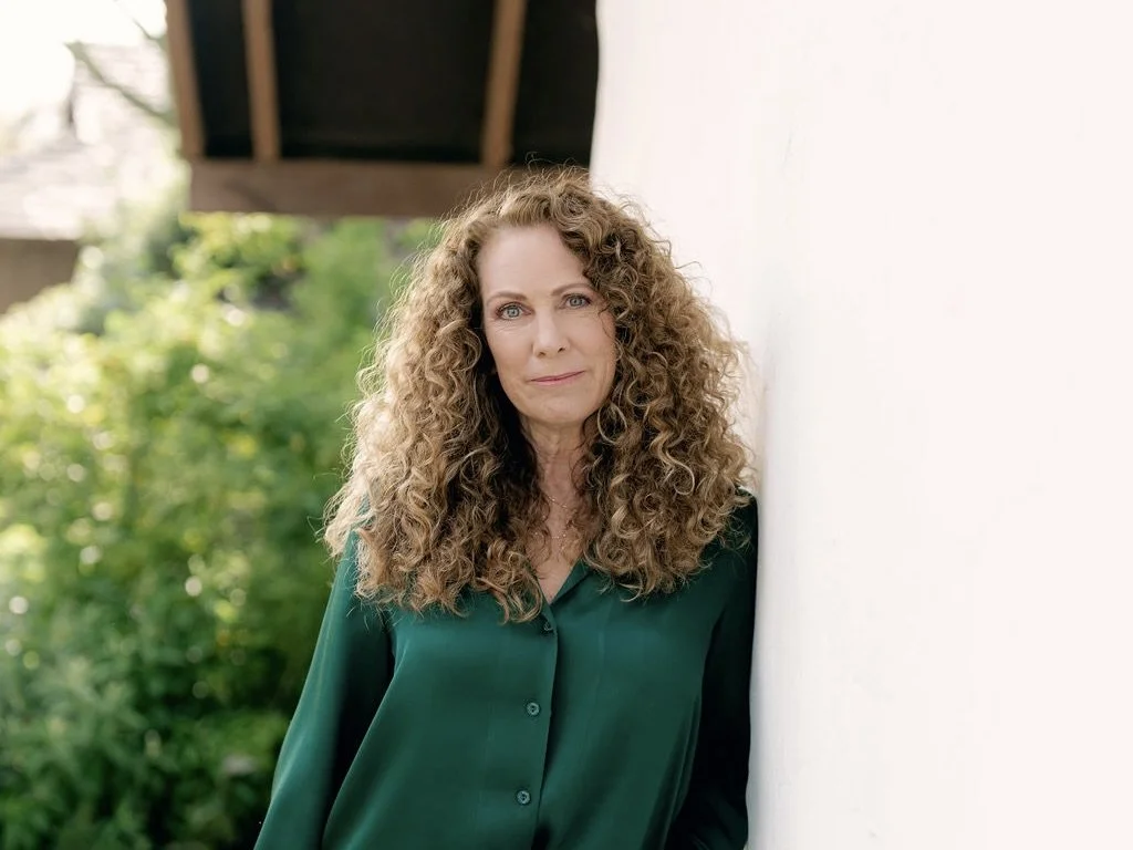 Parenting coach standing calmly outdoors, leaning against a white wall with greenery in the background