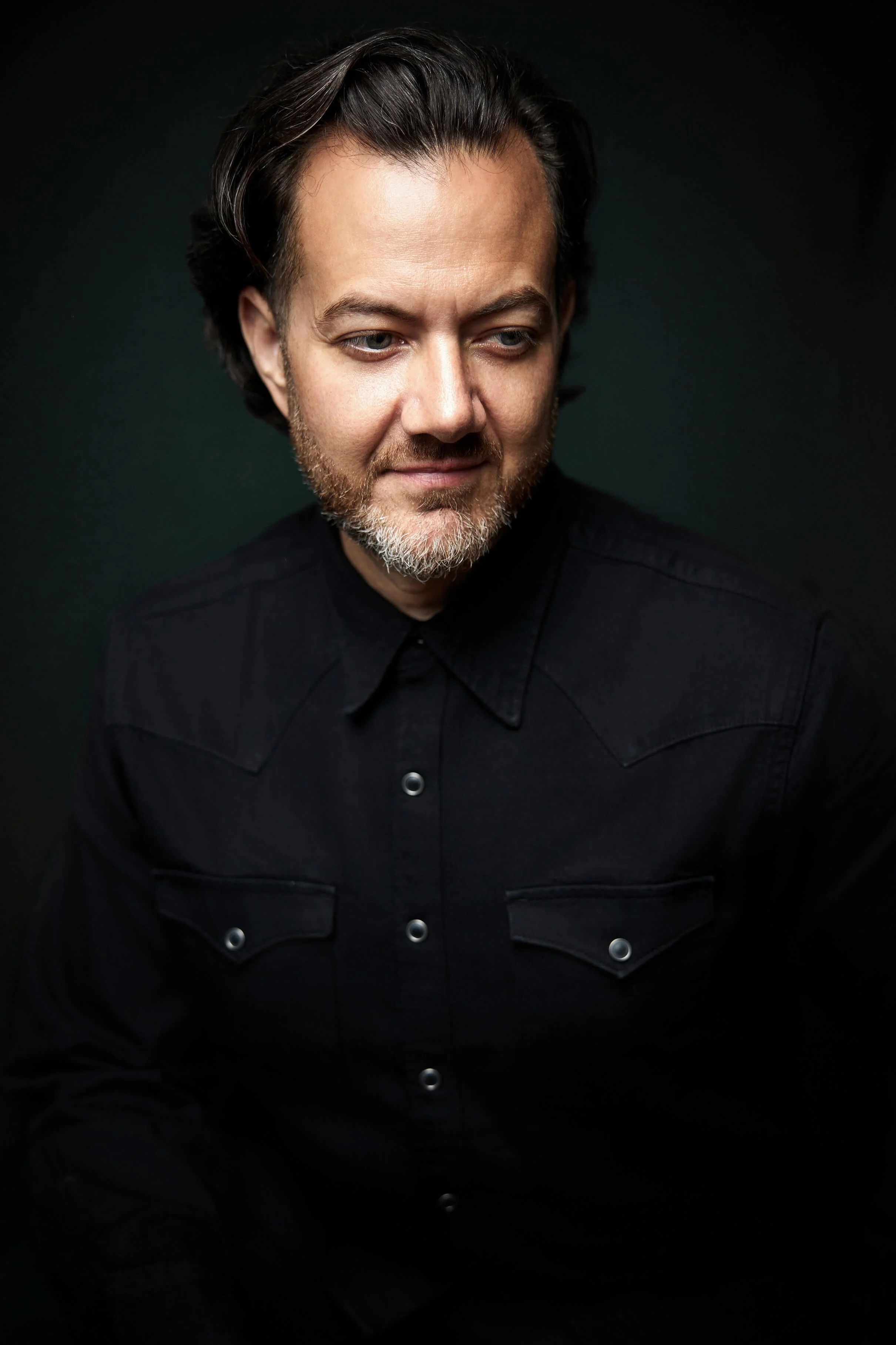 A man with dark hair and a beard, wearing a black shirt, looking slightly to his right against a dark background.