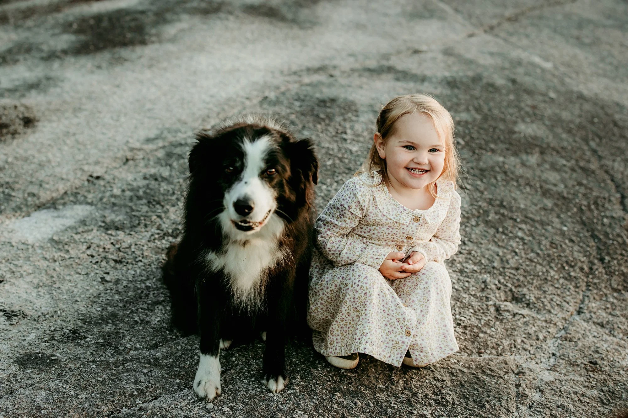A happy young girl with blonde hair and a dog sitting on a gravel surface outdoors.