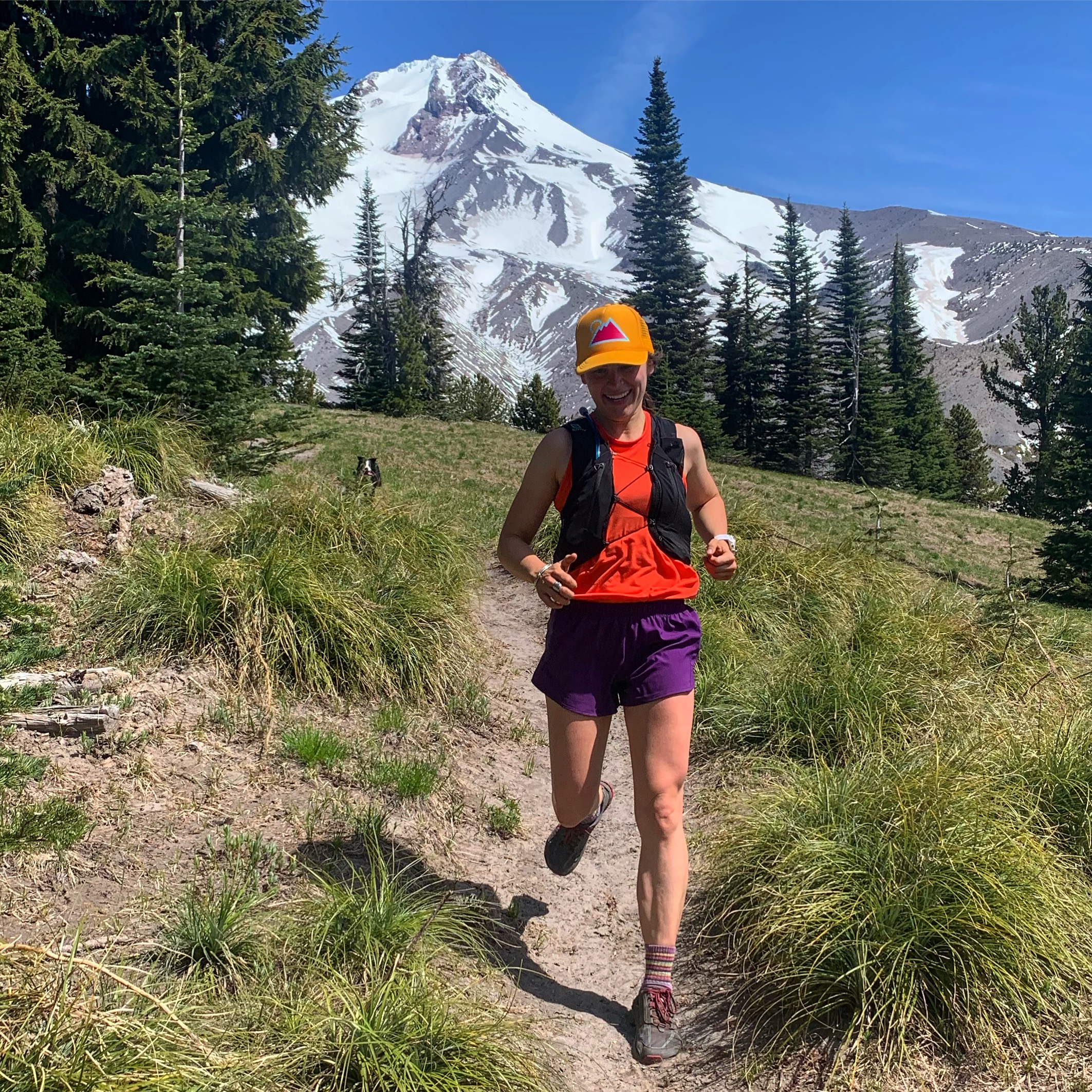 A person trail running on a dirt path in a mountainous forested area with snow-capped peaks in the background.