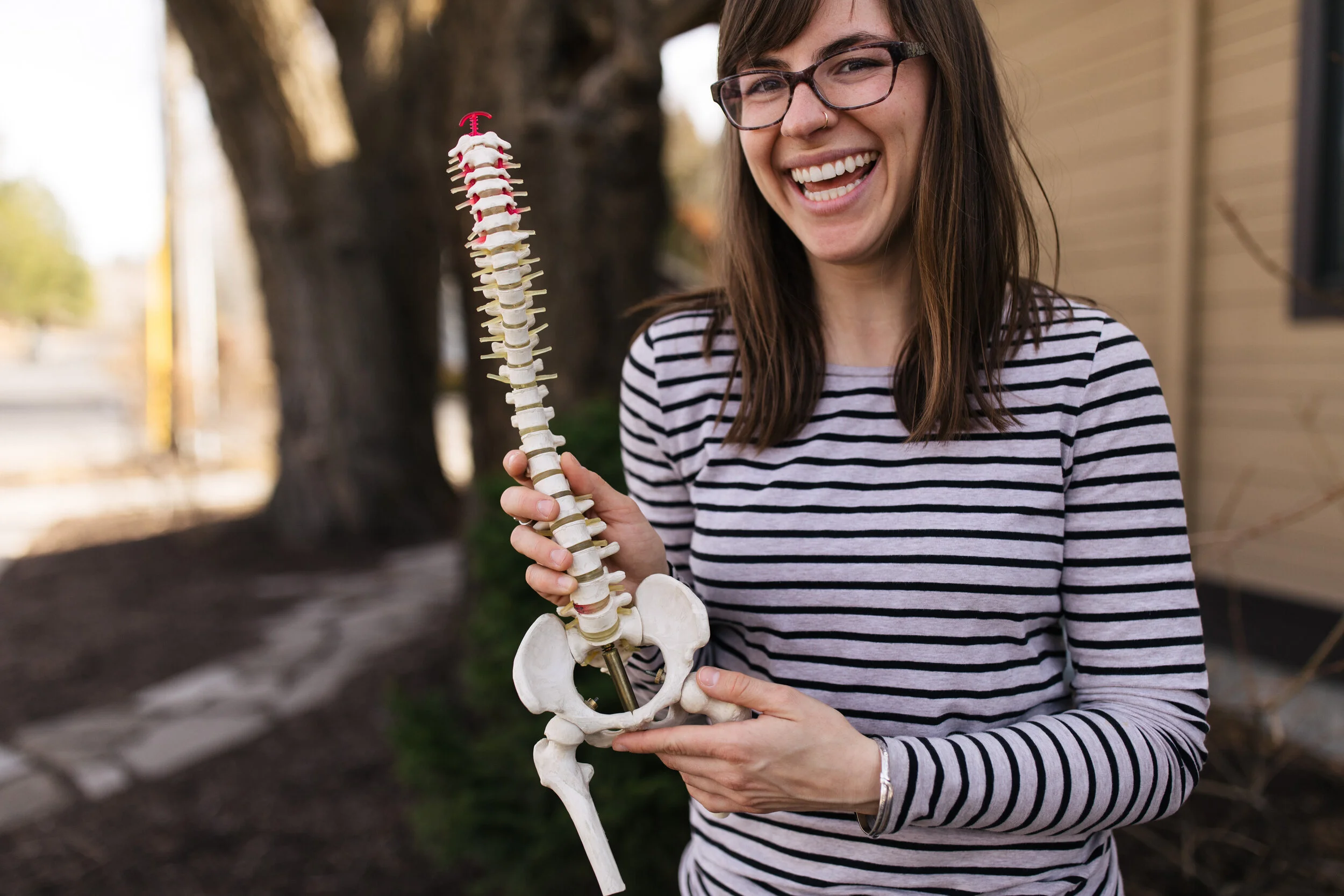 Woman smiling outdoors holding a model of a human spine and pelvis