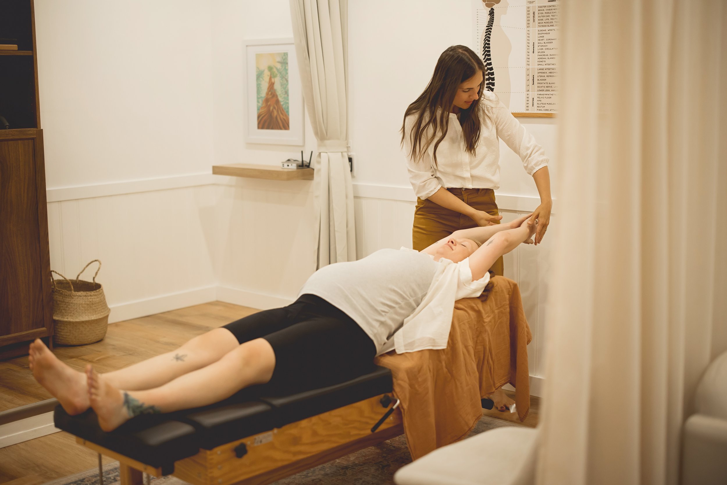 A woman receiving therapy on a massage table from a therapist in a therapy room. The woman is lying on her back with arms extended upwards, and the therapist is holding her arm, demonstrating a stretch or therapy exercise. The room has a framed picture on the wall, a spinal diagram, a basket, and curtains.