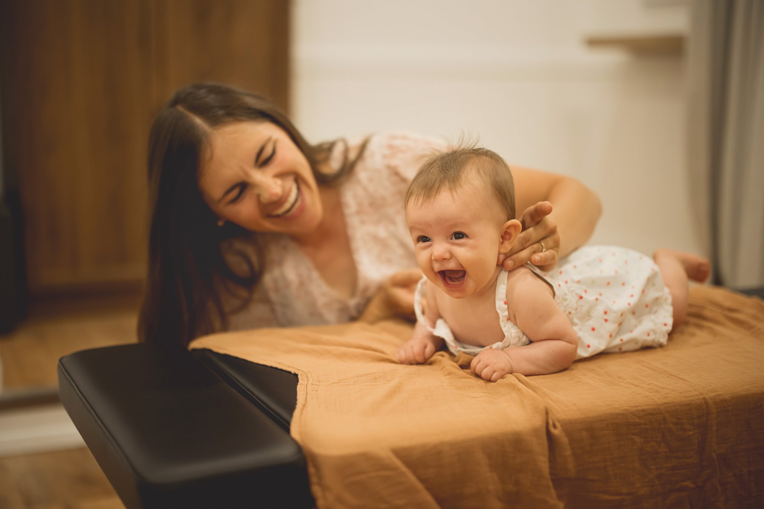 A woman and a baby girl lie on a bed, smiling and laughing together.