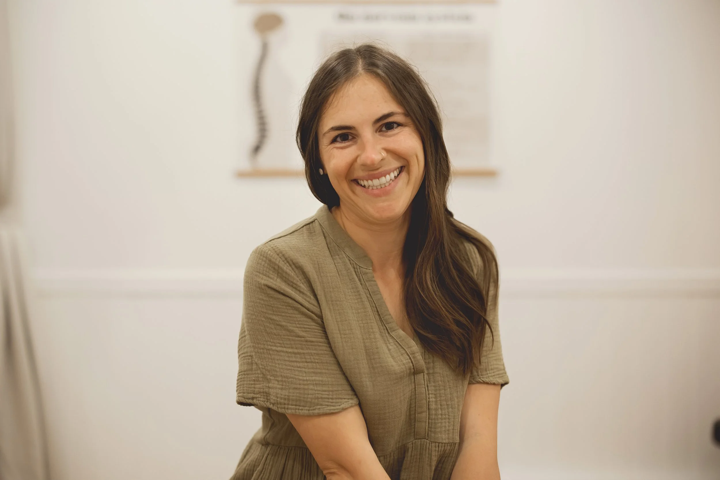 A woman with long brown hair and a tan-colored shirt, smiling at the camera in an indoor setting.