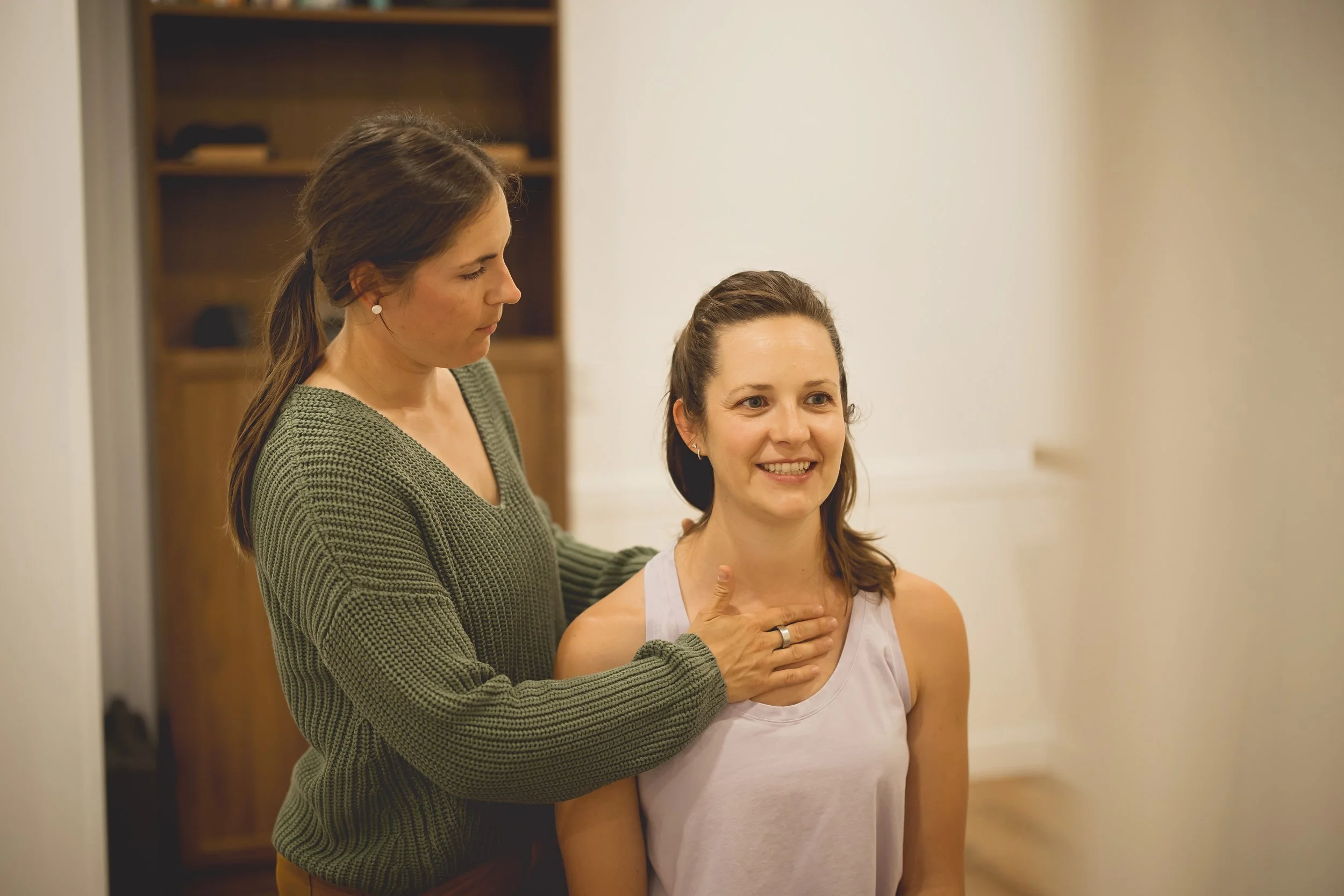 Two women in a home setting, one standing with her hand on the other's shoulder, the other smiling. The woman in front is sitting wearing a white tank top, and the woman behind is wearing a green sweater.