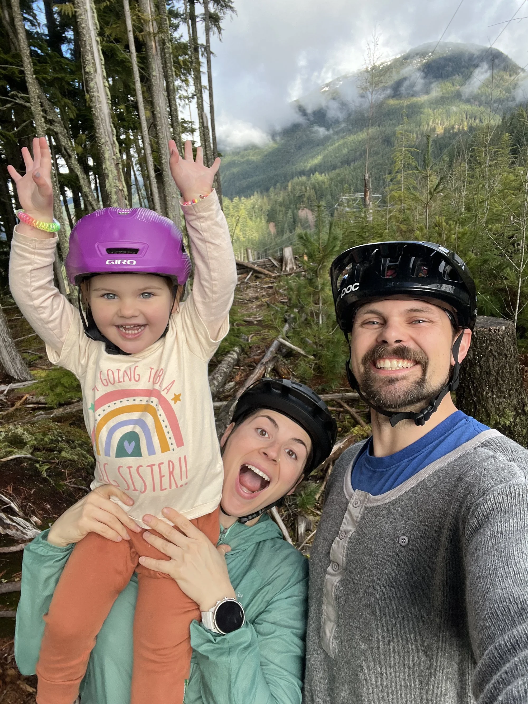 A family of three wearing helmets and outdoor clothing, taking a selfie in a forest with mountains in the background.