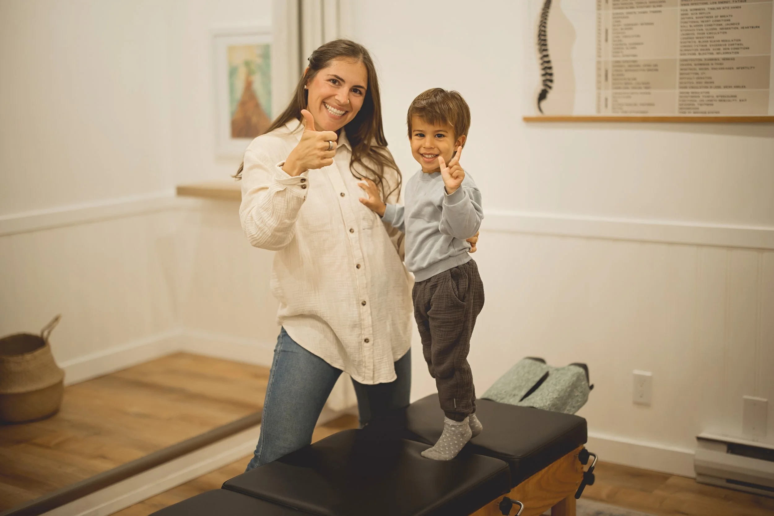 A woman and a young boy standing on an examination table in a medical office, smiling and giving thumbs-up signs.