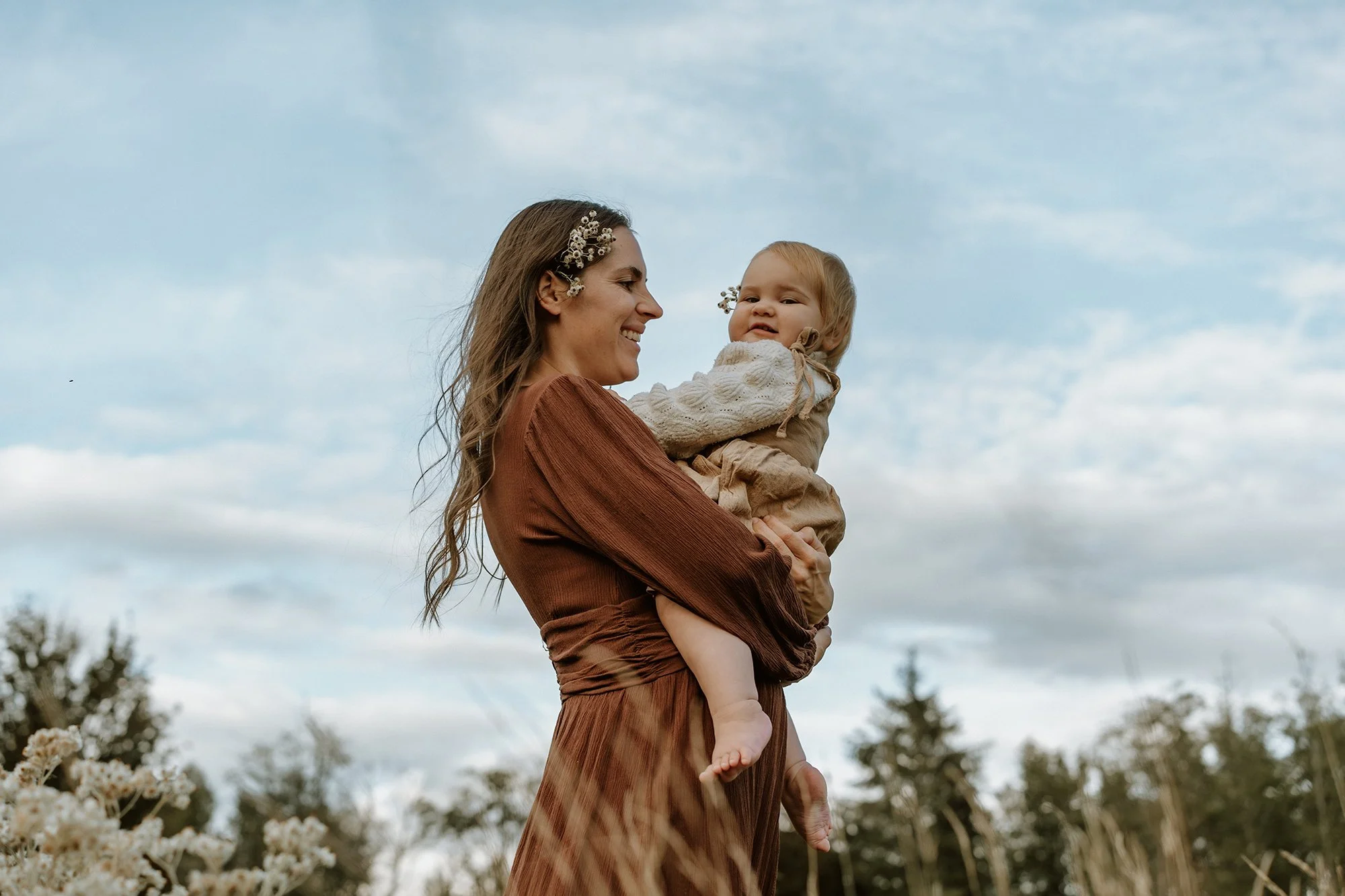 A woman with long wavy hair and a floral hair accessory holding a young child in a field with a cloudy sky in the background.