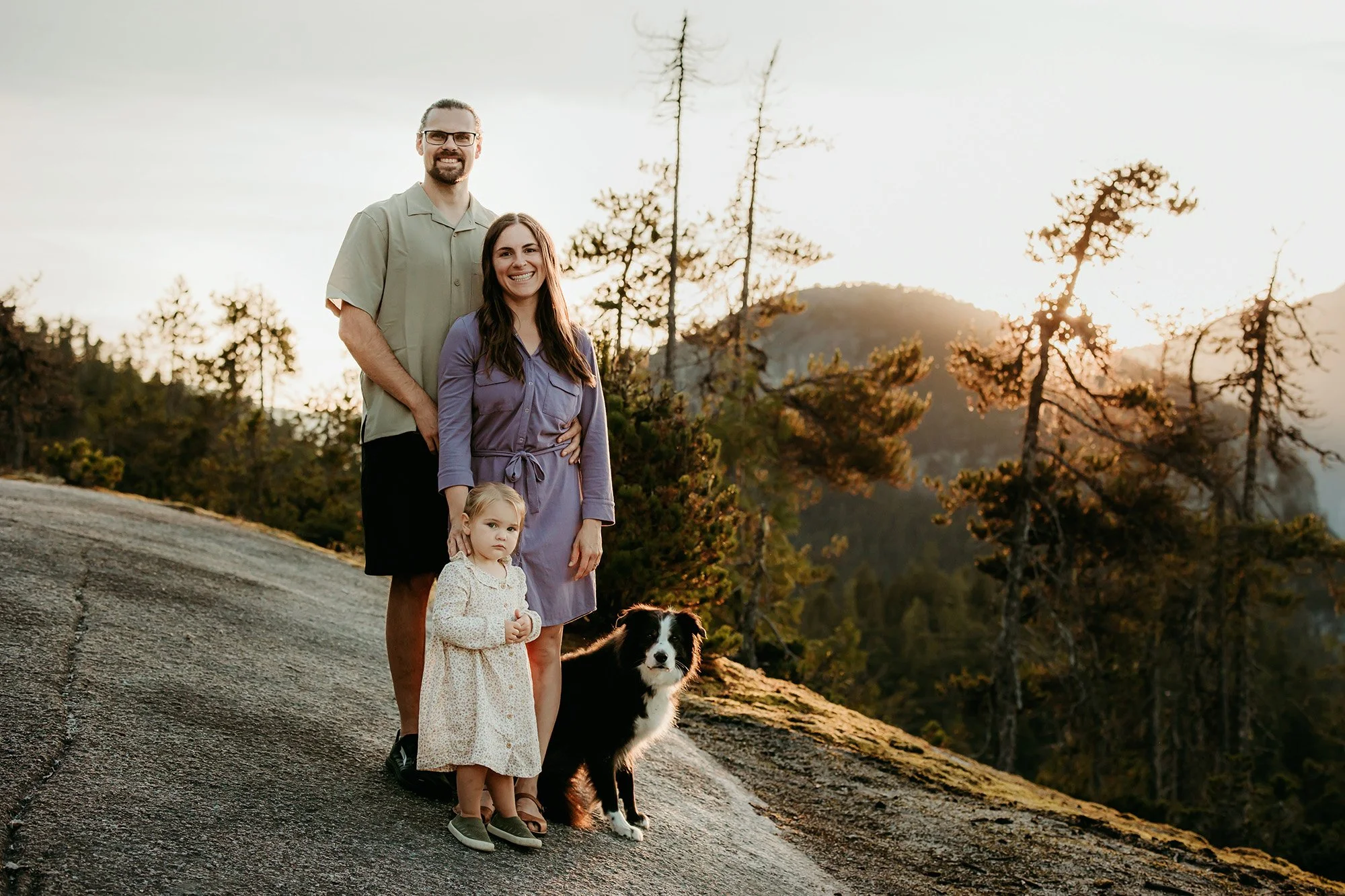 A family of four with a dog standing on a rocky hillside at sunset, surrounded by trees and mountains.
