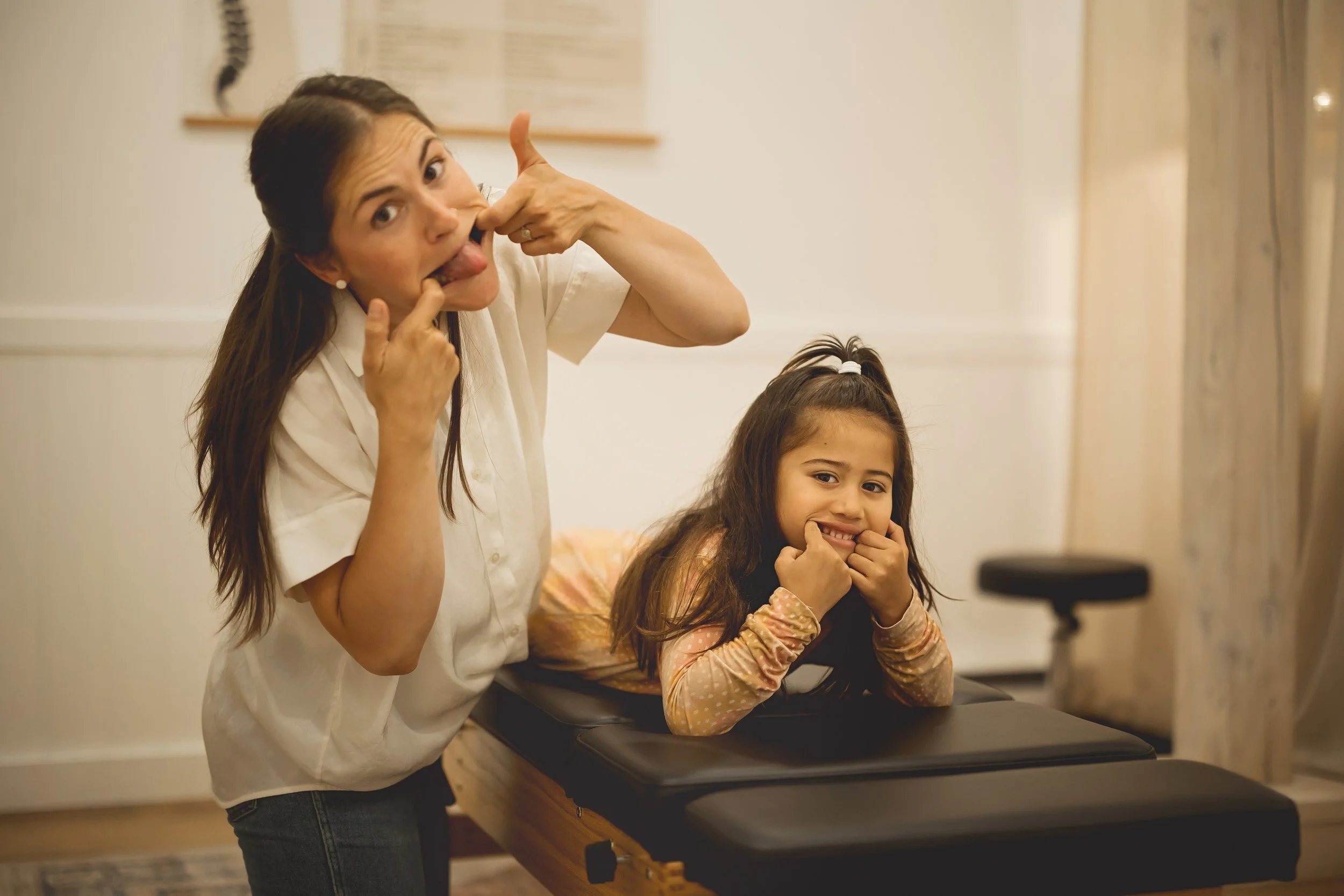 A woman and young girl making funny faces and sticking their tongues out as they pose together in a room with beige walls.
