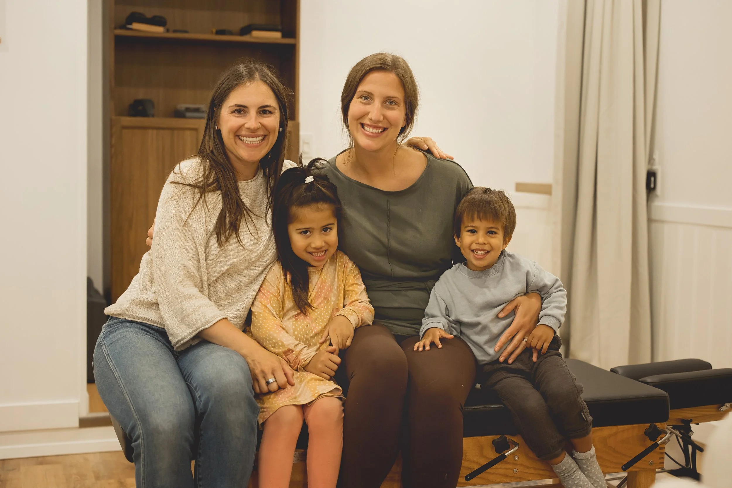 Two women and two children sitting together on a padded bench in a room, smiling at the camera.