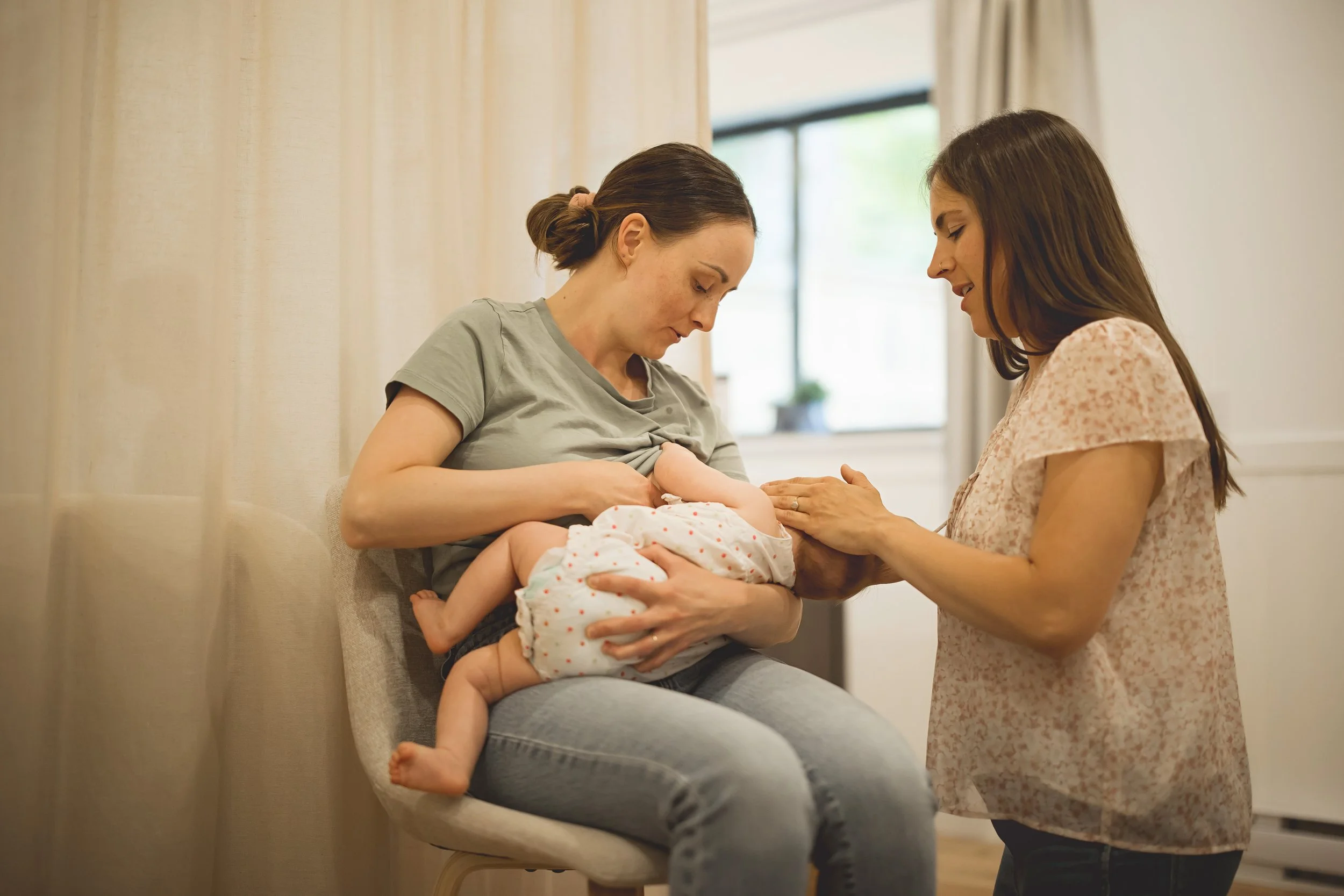 Woman breastfeeding a baby while another woman gently touches the baby's hand in a cozy, well-lit room.