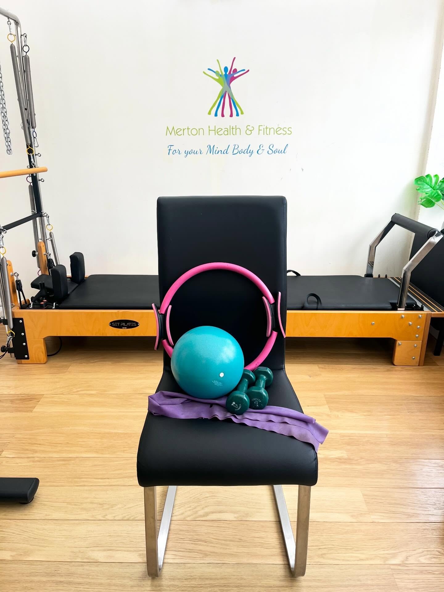 Fitness equipment on a black chair in a fitness studio with a pilates reformer in the background, a teal exercise ball, pink headphones, green dumbbells, and a purple towel.