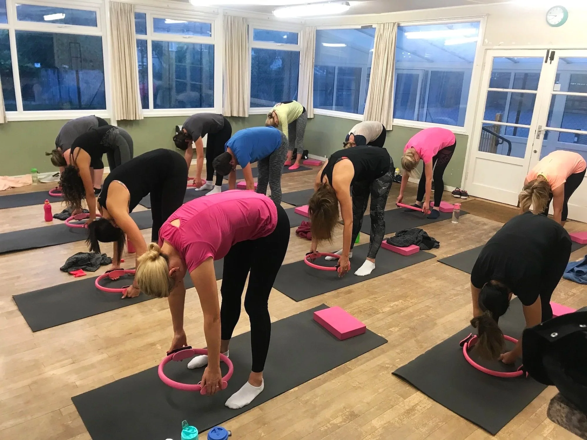 A group of women participating in a Pilates class in a room with large windows, practicing exercises with pink Pilates rings and black mats.