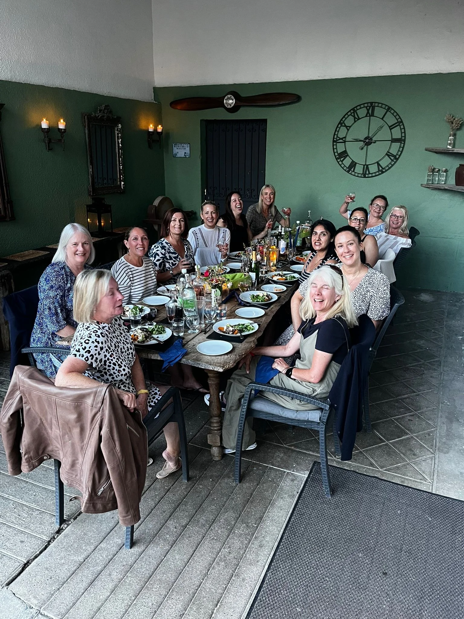 A group of women sitting at a long dining table, enjoying a meal, with some holding glasses raised in celebration.