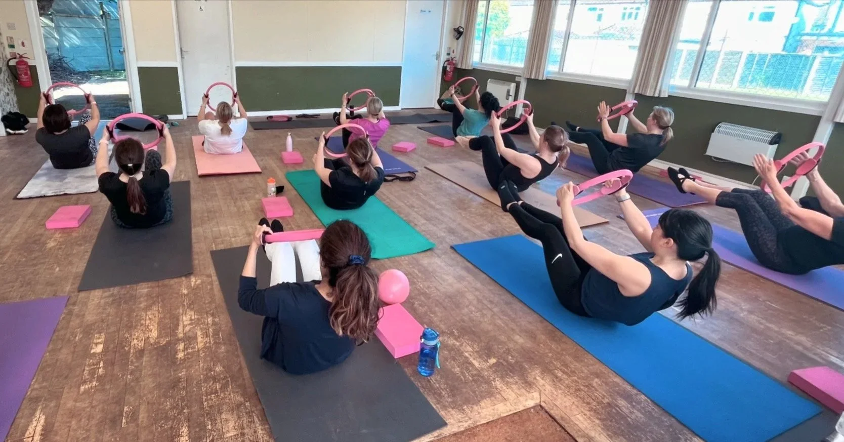 A group of women participating in a fitness class in a studio, sitting on Pilates mats, holding pink fitness rings, with exercise blocks and water bottles nearby.