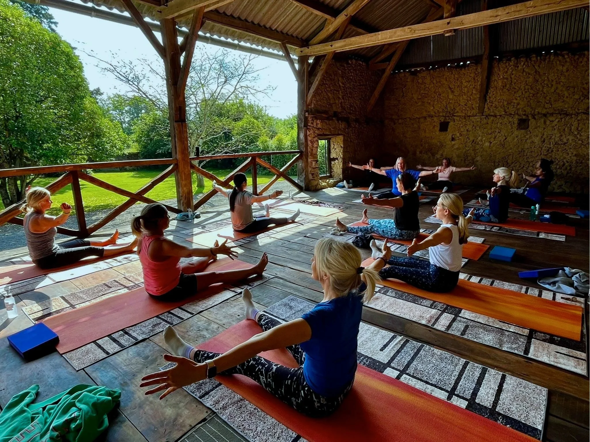 Group of women and children participating in a yoga class outdoors on a wooden deck with green trees and grass in the background.