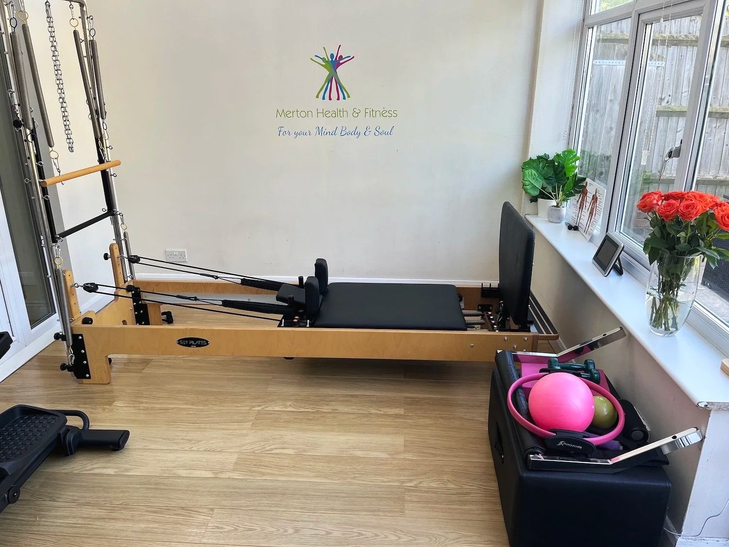 Pilates reformer machine in a bright room with large windows, a potted plant, a vase of red roses, and fitness equipment including a pink ball and resistance bands.