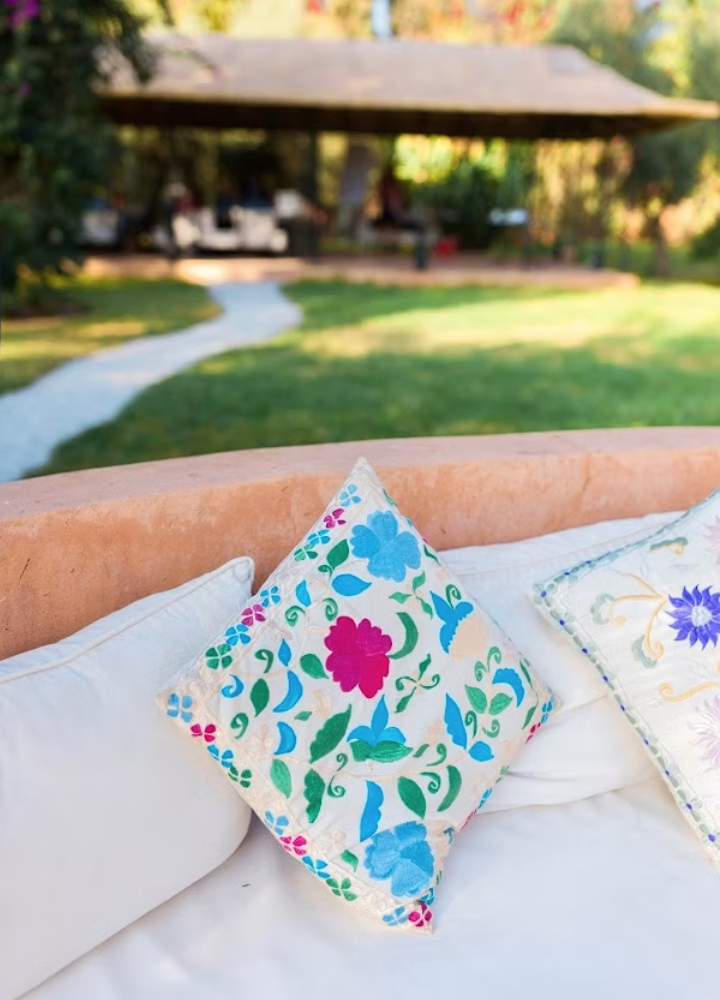 Close-up of decorative pillows with floral embroidery on a white outdoor couch. In the background, there's a pavilion, grass lawn, and a pathway.