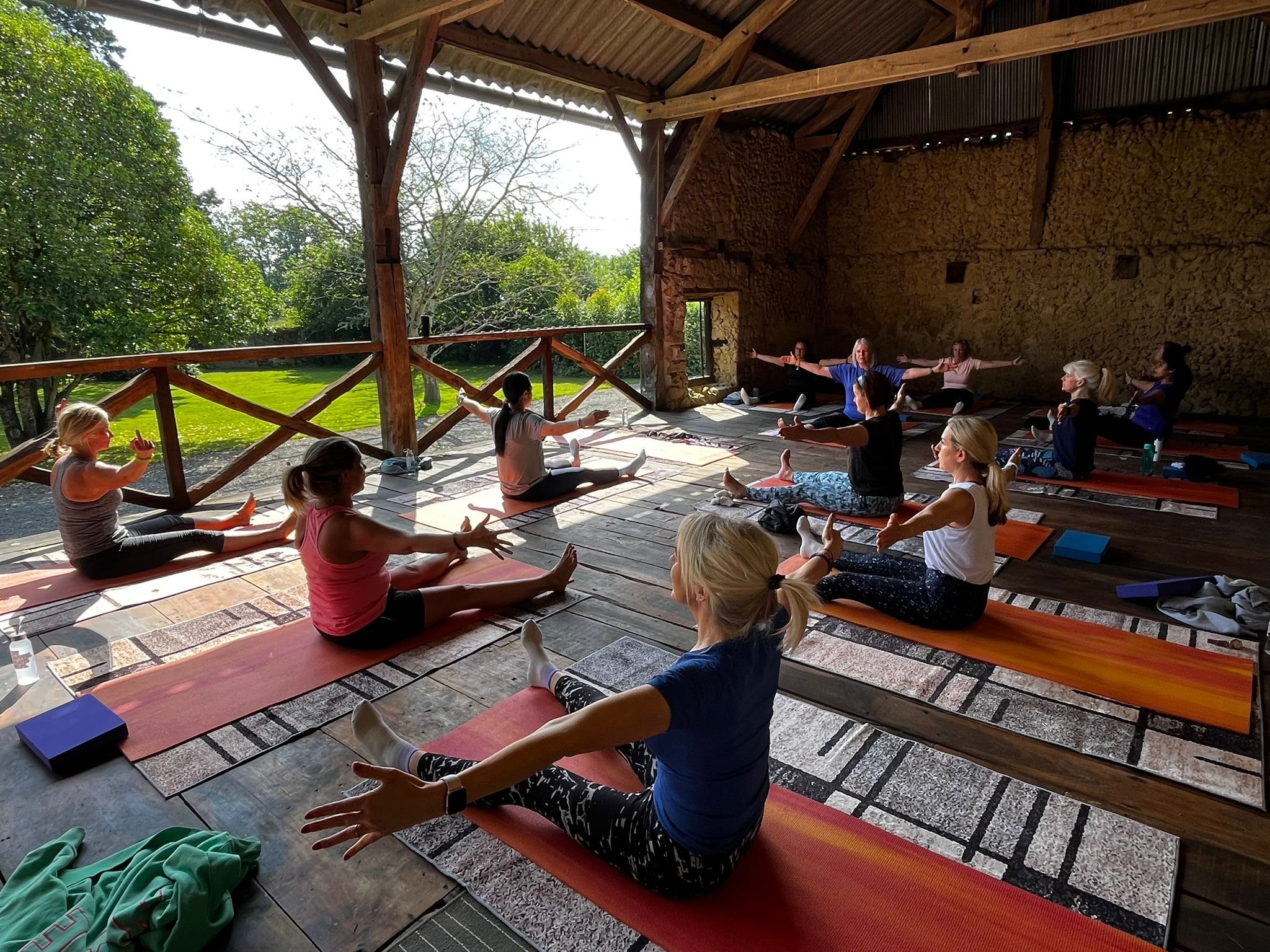 A group of women practicing Pilates at a Pilates retreat in South West France with an open side overlooking green trees and grass. The women are seated on mats, stretching their arms and legs, with some looking towards the instructor at the front.