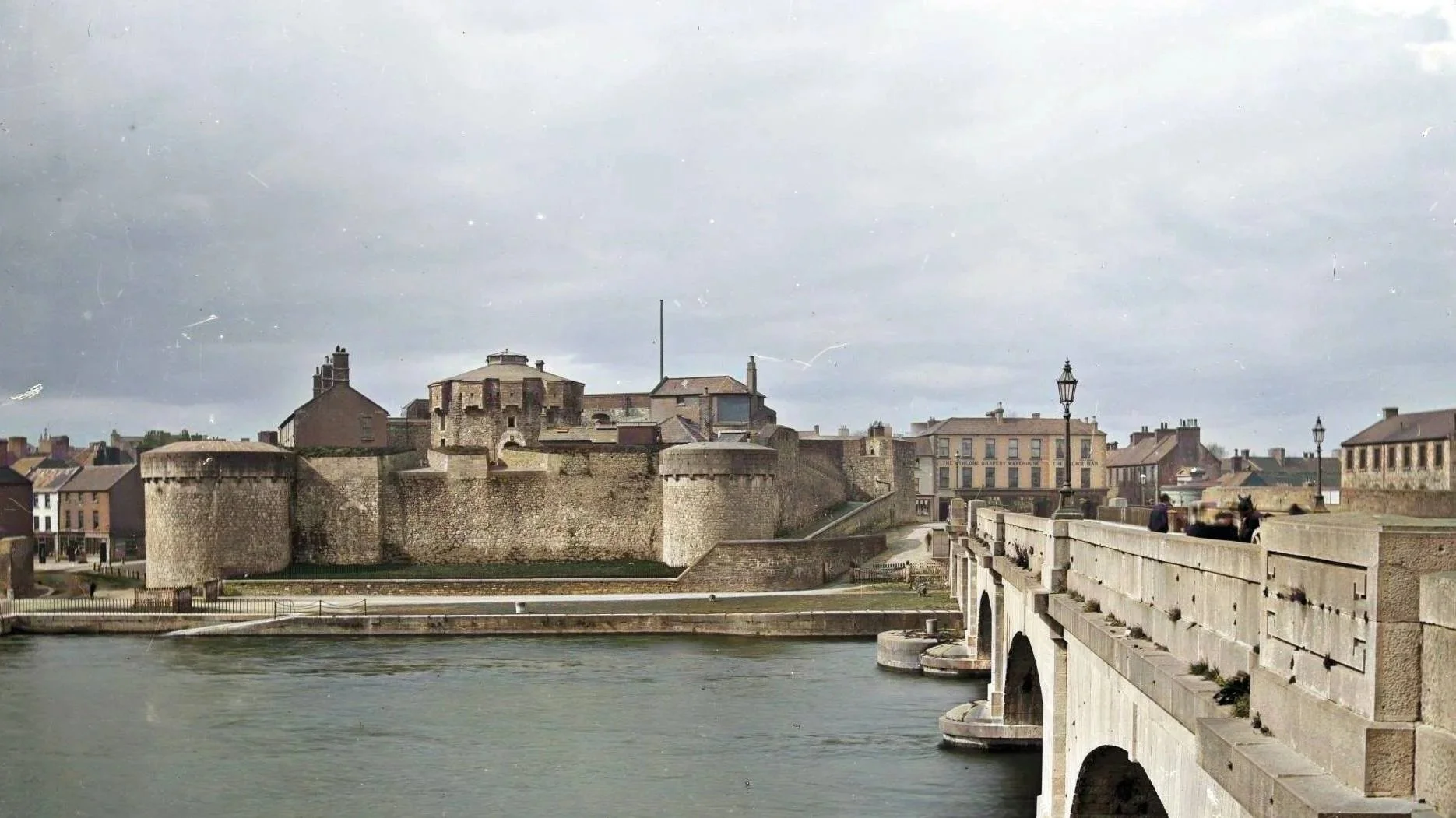 A view of a historic stone castle with rounded towers, surrounded by a river. There is a stone bridge in the foreground with lamp posts and a few people walking across.