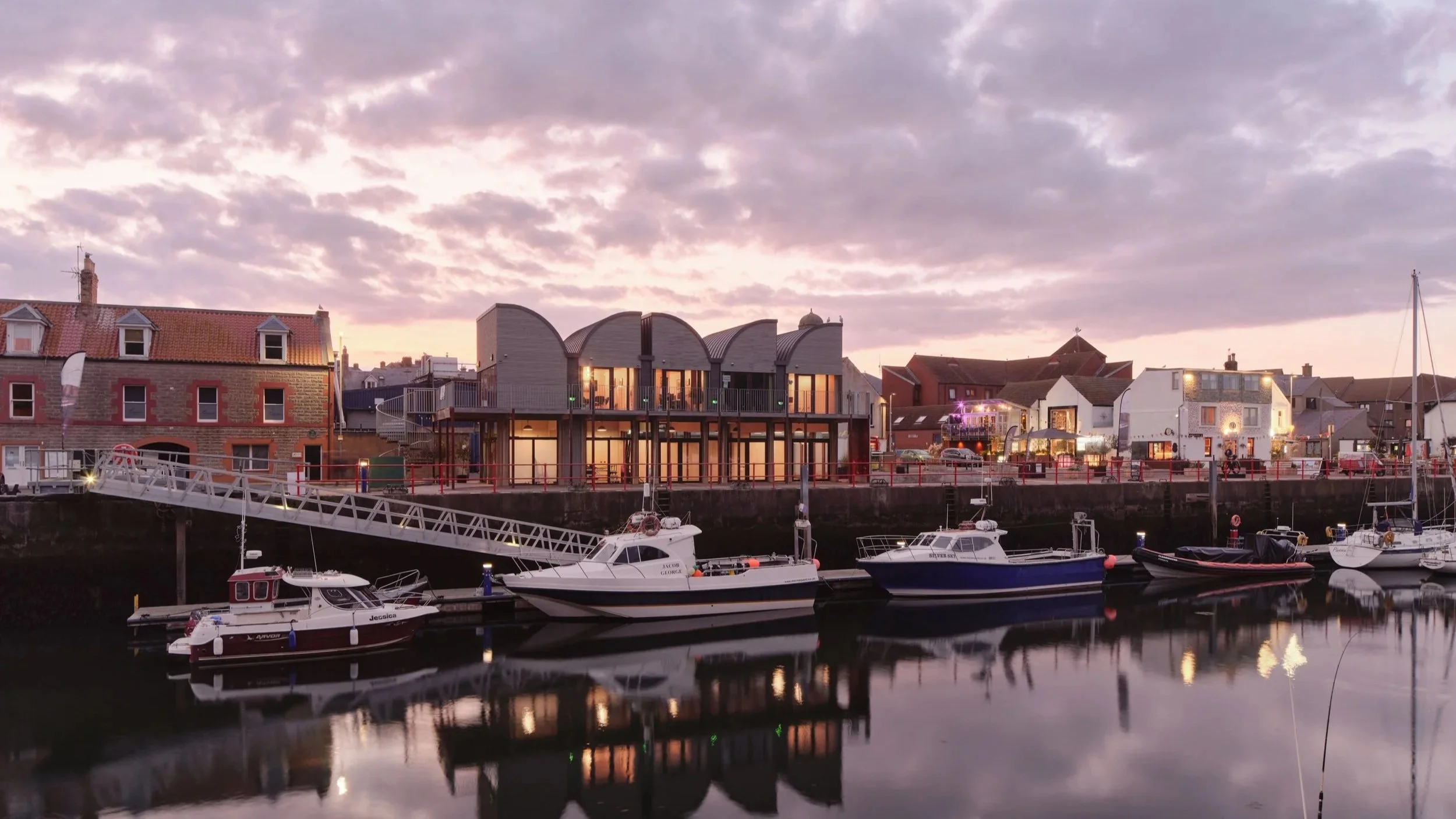 Harbor with docked boats in front of a row of buildings under a cloudy sky at sunset.
