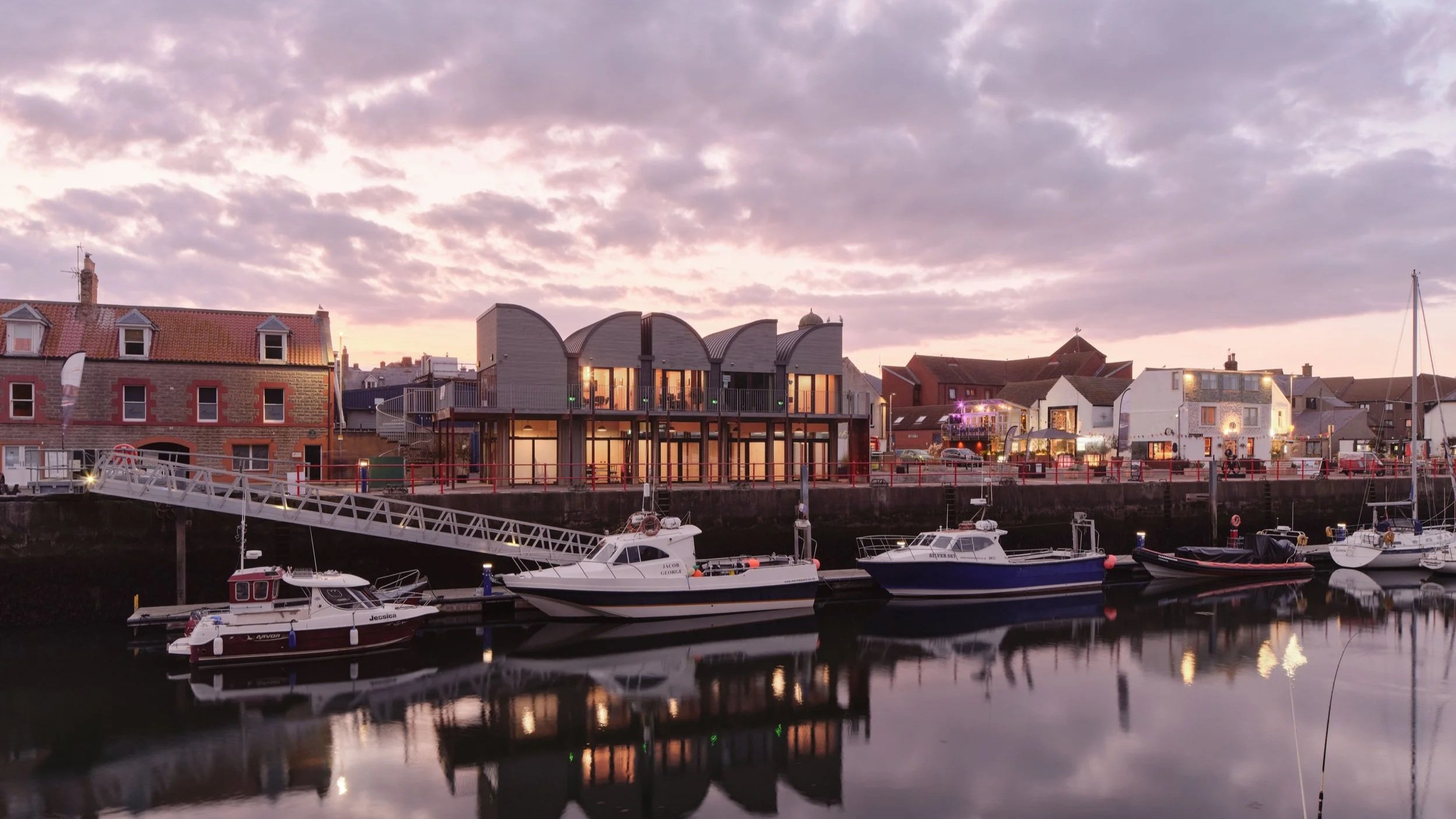 Galmstrup Architects — Harbor with docked boats in front of a row of buildings under a cloudy sky at sunset.