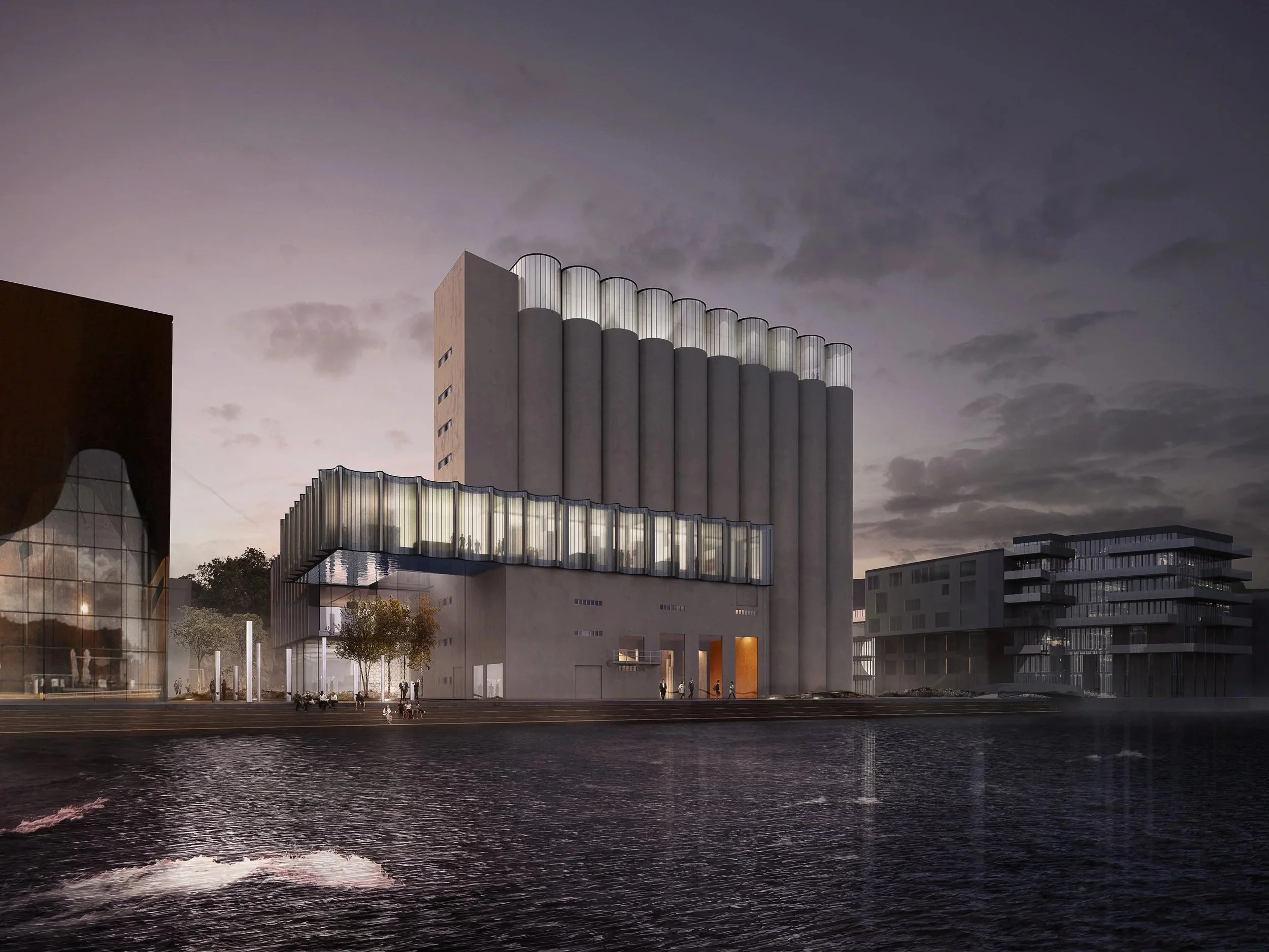 Galmstrup Architects — Nighttime view of a modern architectural building with cylindrical structures on top, glass-enclosed sections, and exterior lighting, situated by a bo