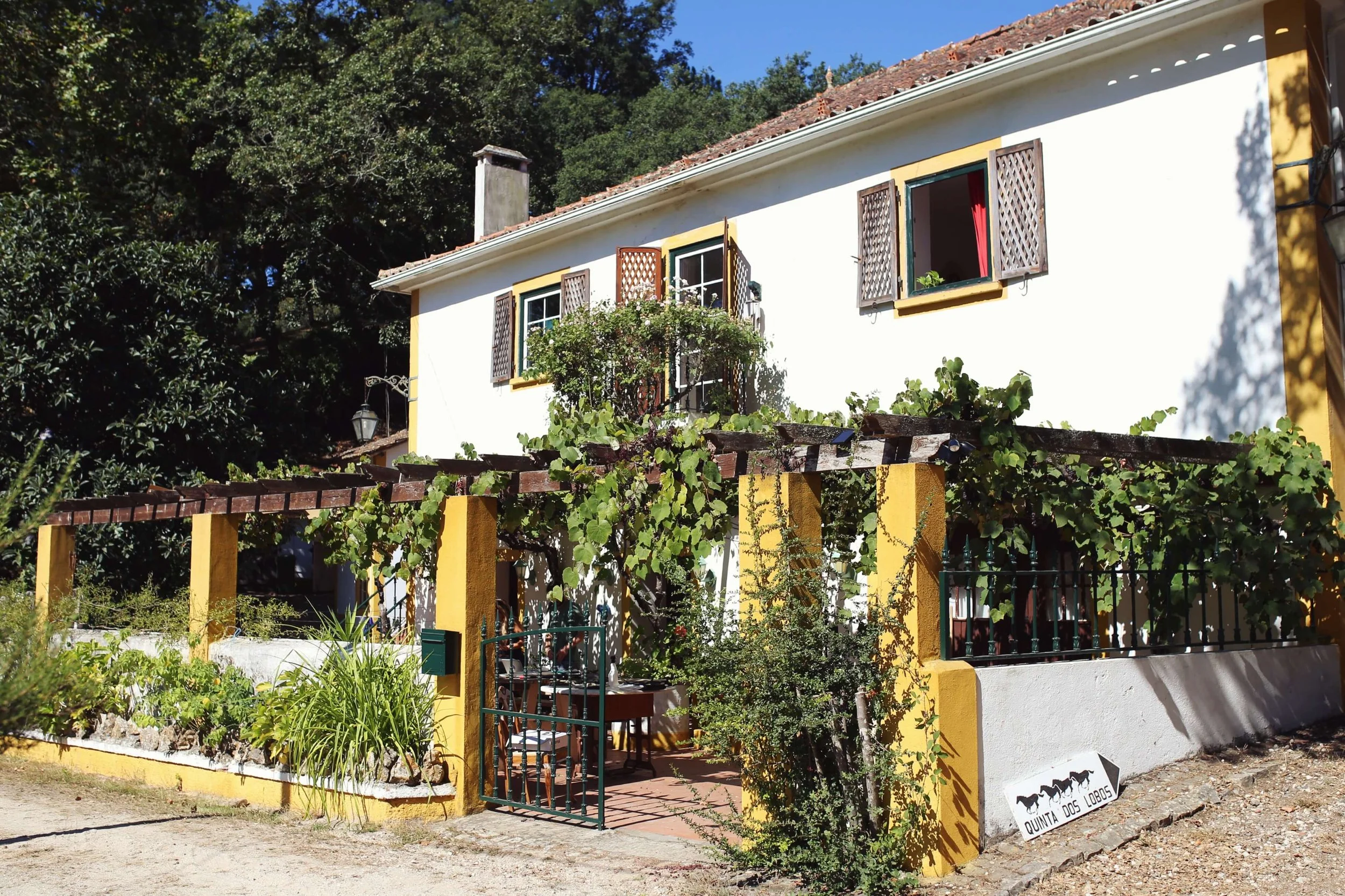 A yellow and white two-story house with open windows, surrounded by greenery and vines, with a sign reading 'Quinta dos Lobos' outside.