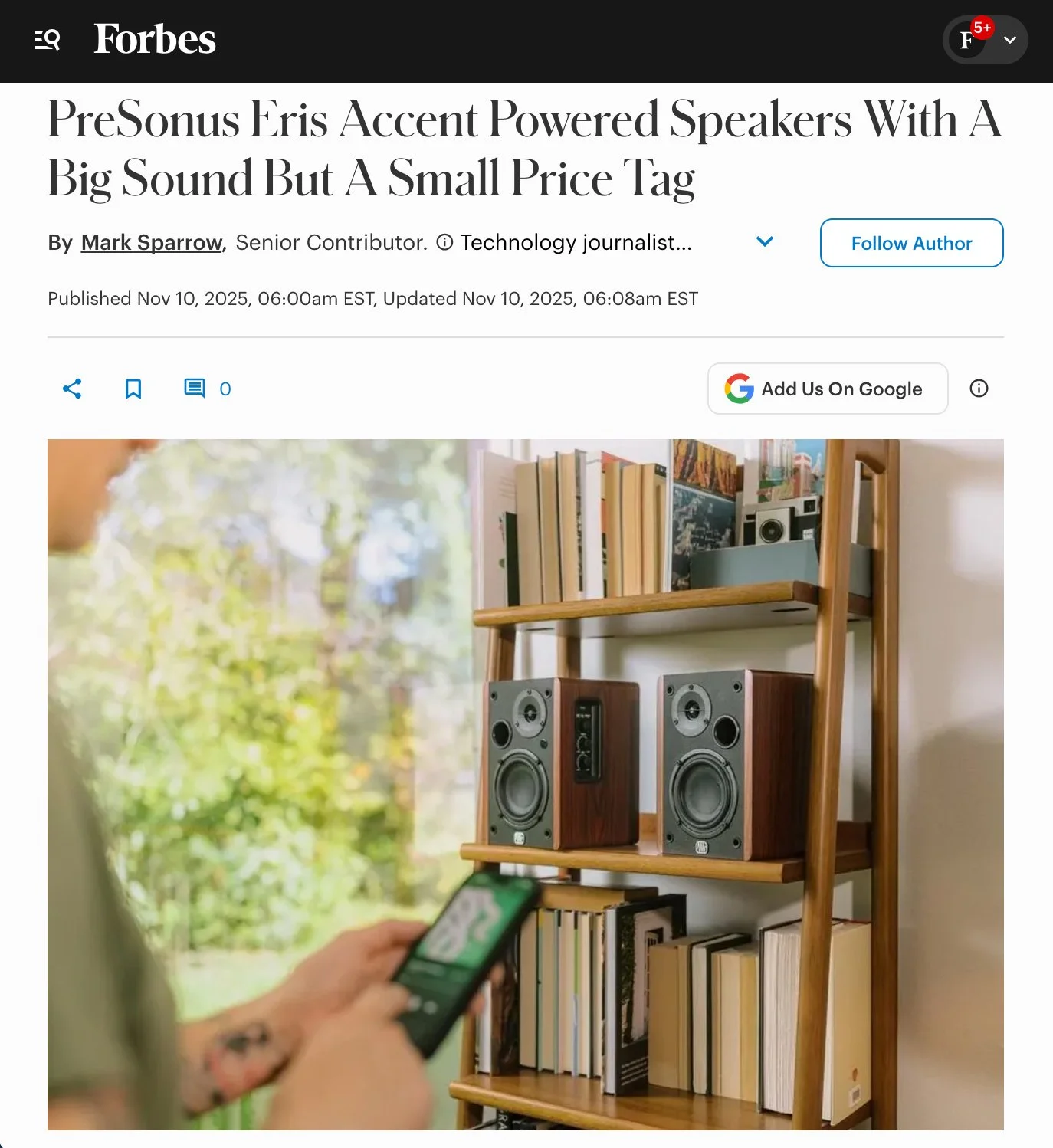 Photo of a wooden bookshelf with books and speakers, with a blurred background of trees visible through a window. A person's hand holding a phone is in the foreground.