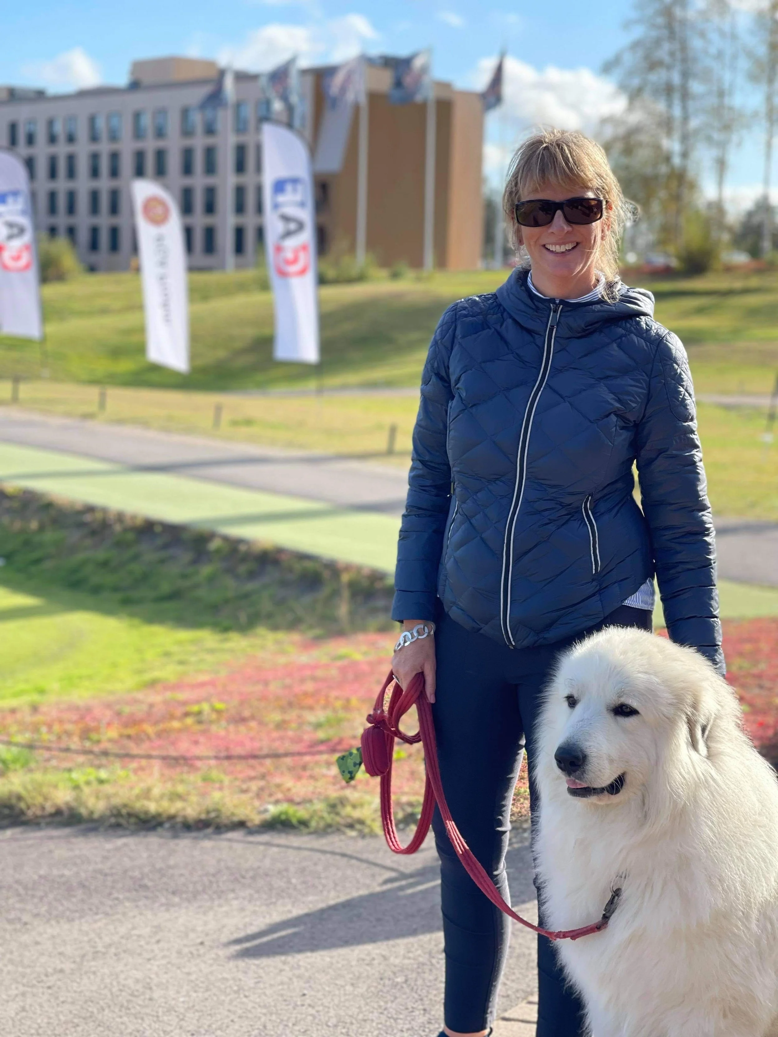 A woman smiling in sunglasses holding a red leash attached to a large white retriever dog outdoors on a sunny day. In the background, there are flags and a building.