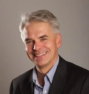 A smiling middle-aged man with gray hair, wearing a dark blazer and light blue shirt against a neutral background.