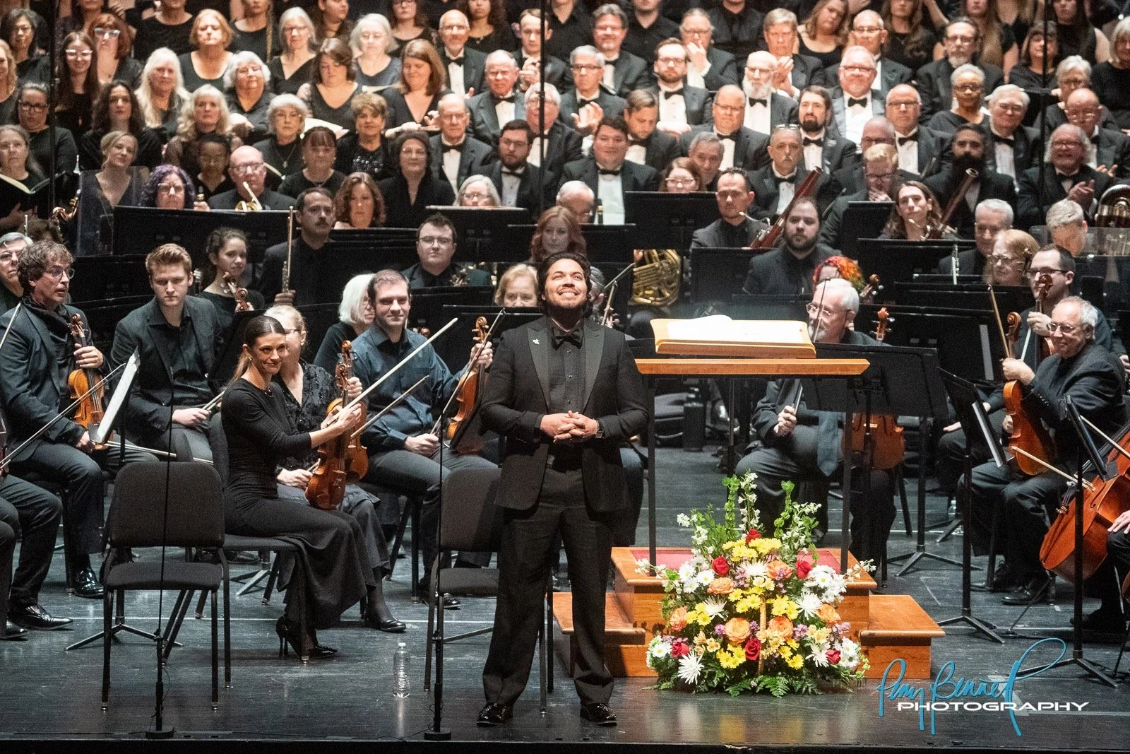 baritone Ethan Vincent in a black tuxedo stands on stage with a large orchestra behind him, all in formal attire, with a floral arrangement in front. The audience is visible in the background, seated and clapping.