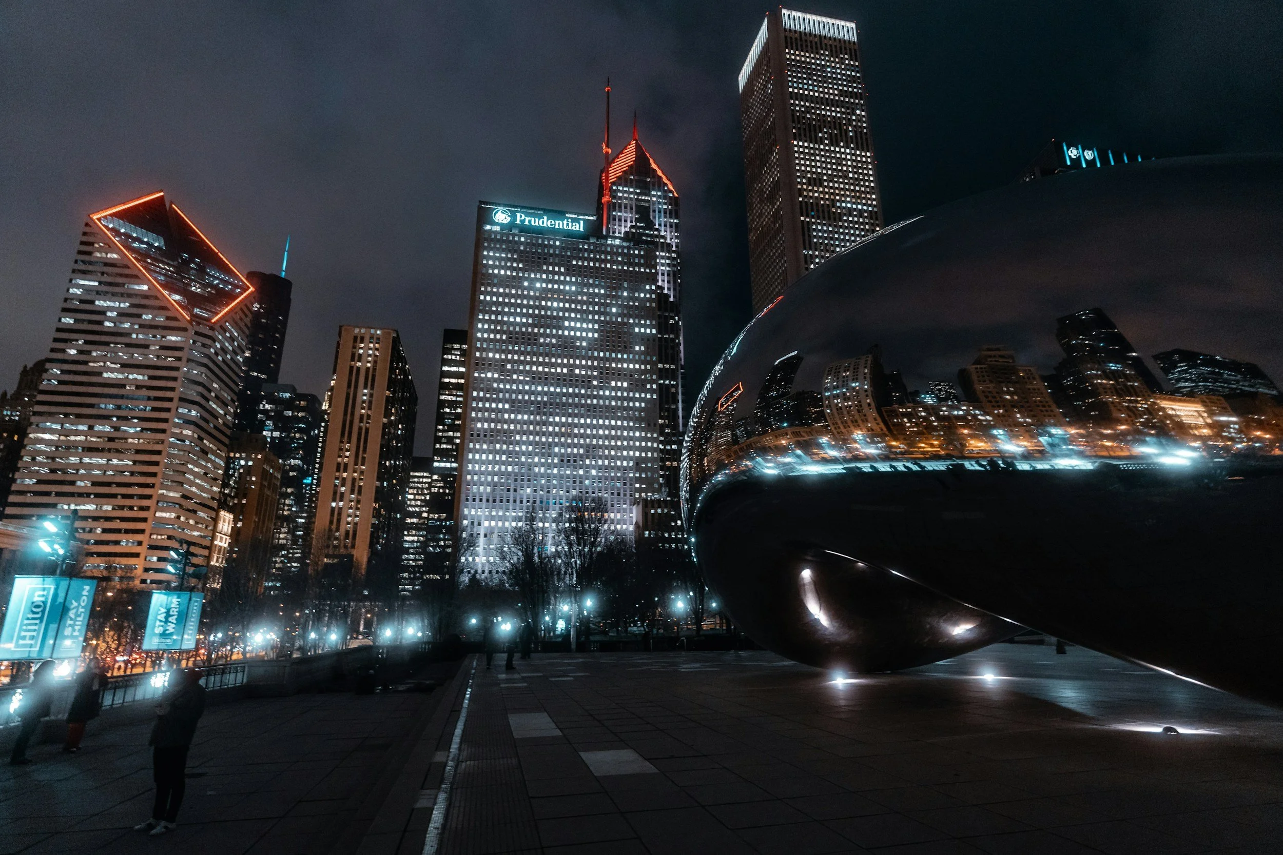 Long exposure photo of light trails from moving vehicles on a curved road near a modern building at night, with trees and a body of water in the background.