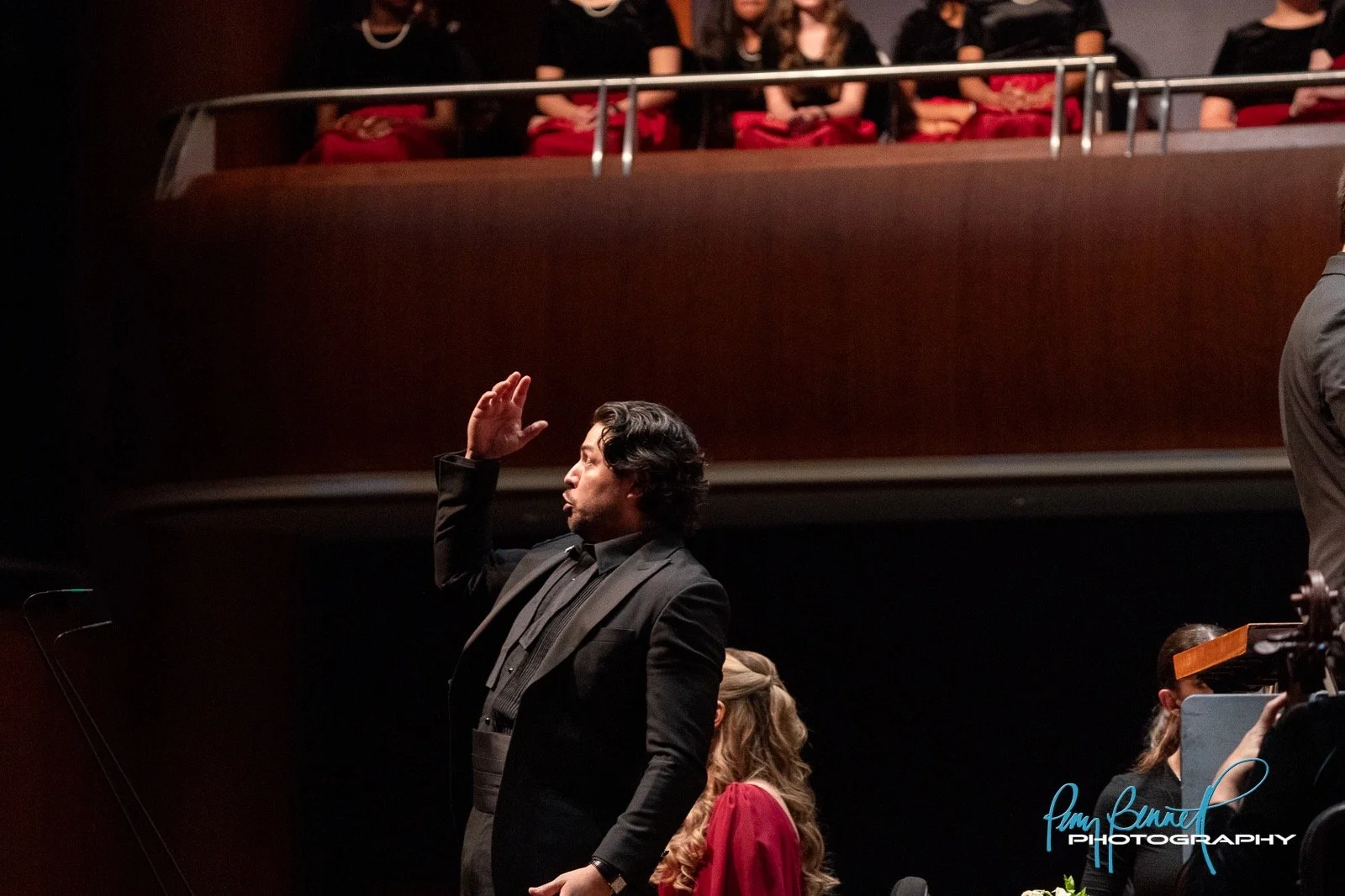 Ethan Vincent baritone in a black suit and black shirt with long wavy black hair, saluting with his right hand in a courtroom or formal setting, with women seated behind him and others in the background.