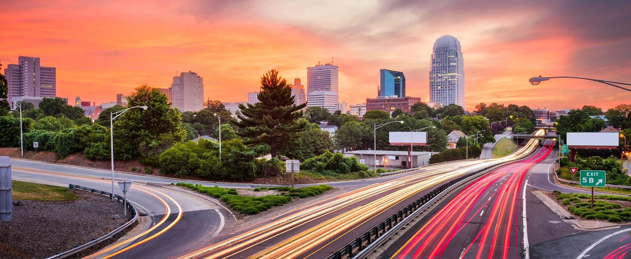 Blurred cityscape with sunset sky, buildings in the background, and a road with cars in the foreground.