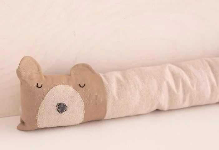 A beige plush bear head pillow attached to a long, cylindrical body pillow with white fabric, resting against a white wall.