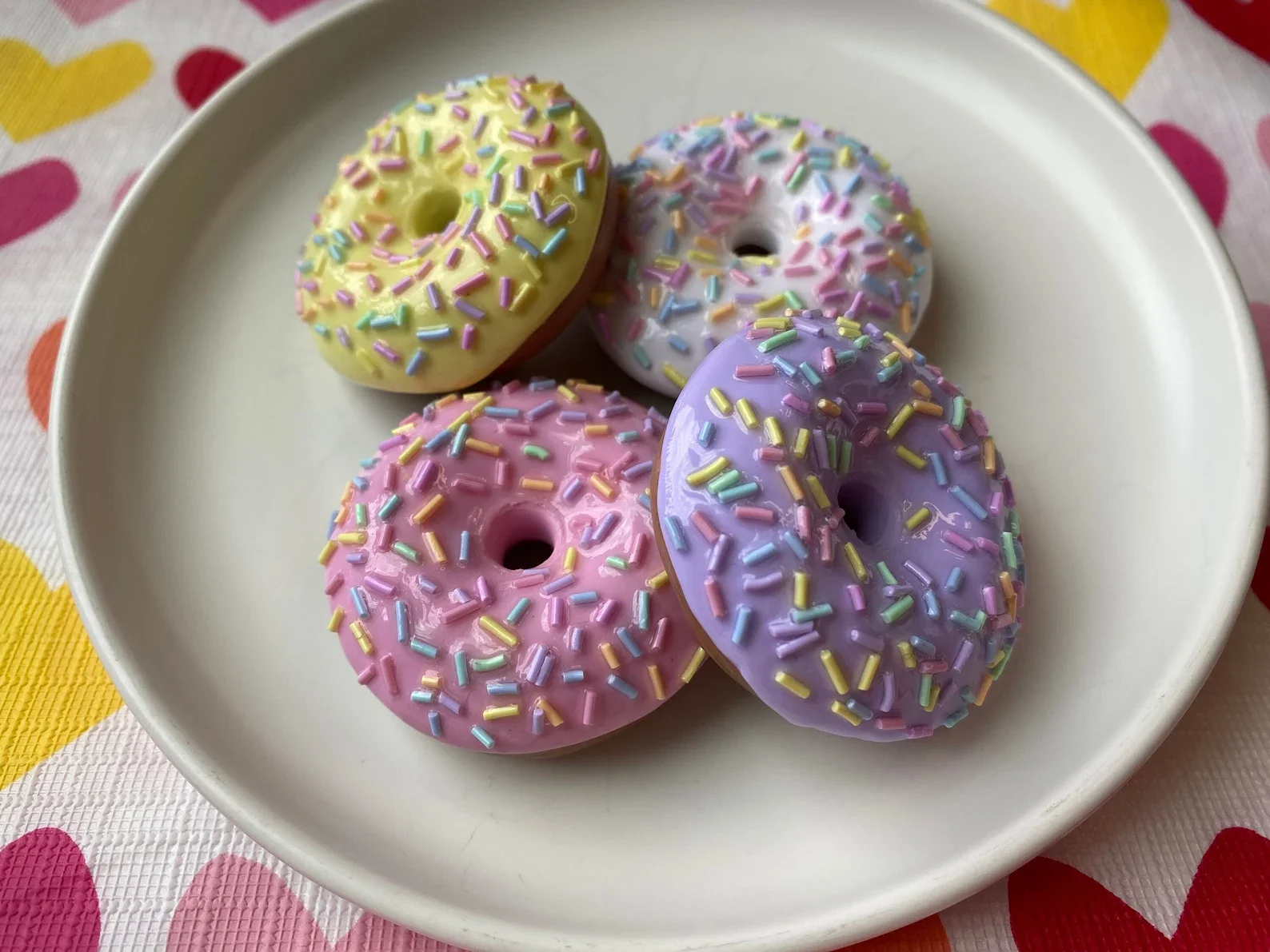 Four colorful donuts with sprinkles on a white plate, set on a heart-patterned tablecloth.