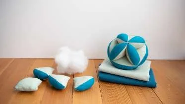 Stacked blue and white cushions on the floor with a geometric plush pillow, some stuffed toys, and a white fluffy object nearby, against a plain white wall.