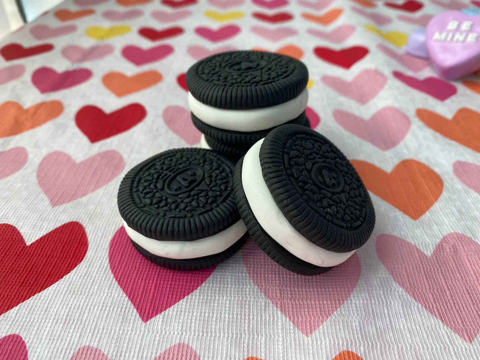 Stack of black-and-white sandwich cookies with an owl design on top, placed on a pink, red, orange, and purple heart-patterned tablecloth.
