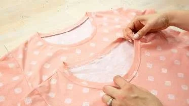 A person is folding a pink and white checkered t-shirt on a light-colored surface.