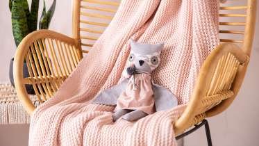 A stuffed raccoon toy dressed in pink clothing sitting on a pink blanket draped over a wooden chair with rattan backrest.