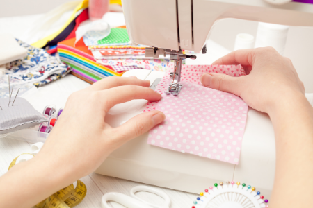 Person sewing pink polka dot fabric on a sewing machine surrounded by colorful fabric and sewing supplies.