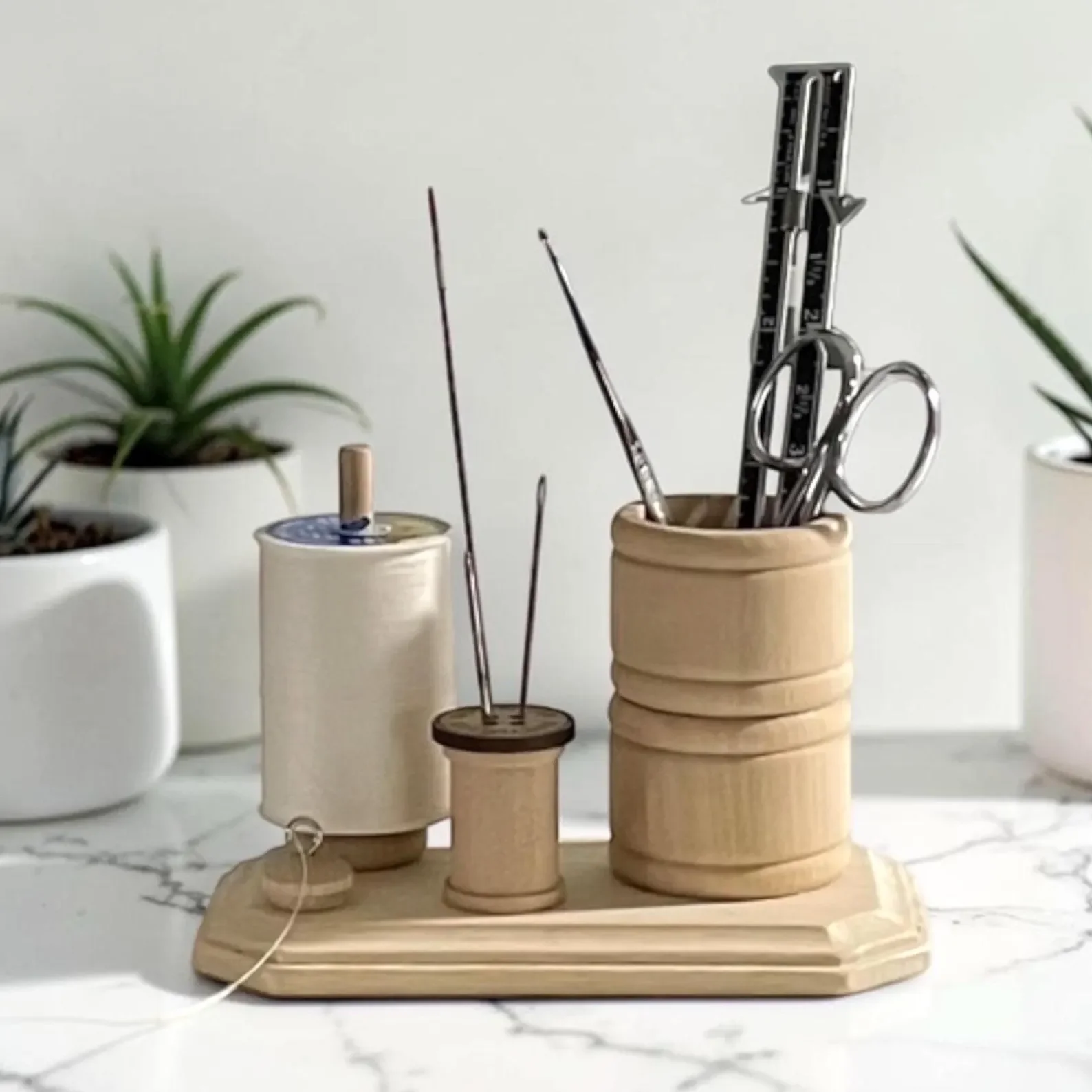 A wooden organizer with various tools, including scissors, a ruler, and a needle, on a marble countertop. Two potted plants are in the background.