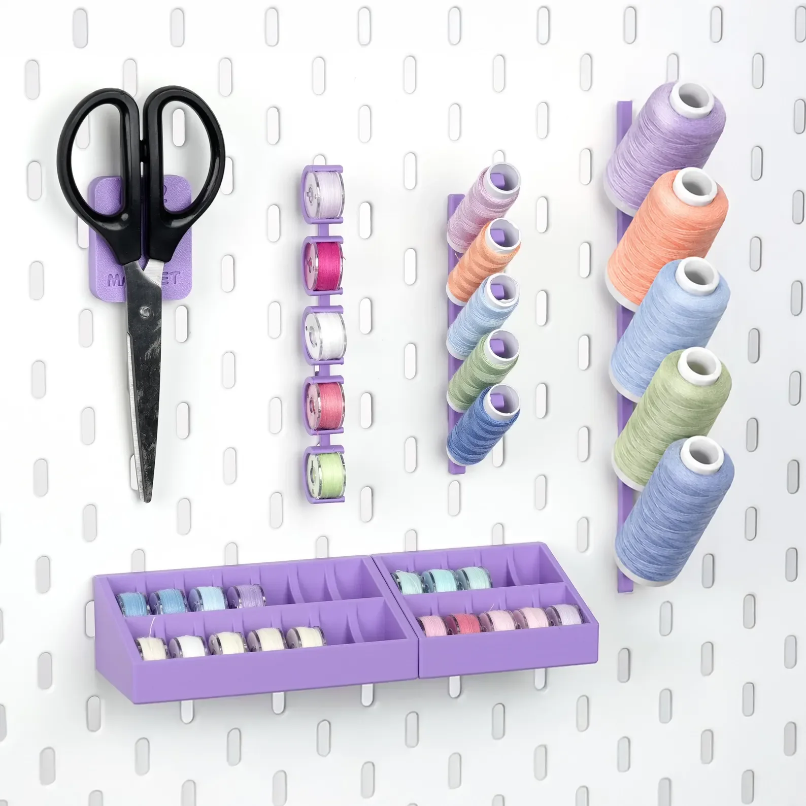 A collection of sewing supplies on a white pegboard. There is a pair of black scissors with purple handles, a purple and pink thread storage bobbin holder, and multiple spools of pastel-colored thread. Additionally, a purple compartment holds small spools of thread.