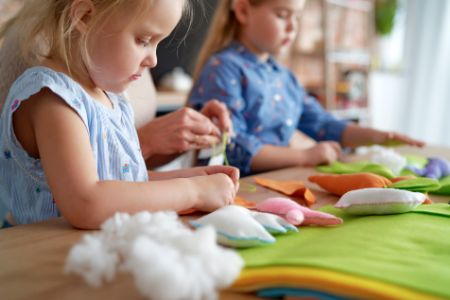 Two young girls are sitting at a table working on a craft project with colorful fabric, stuffing, and sewing supplies.